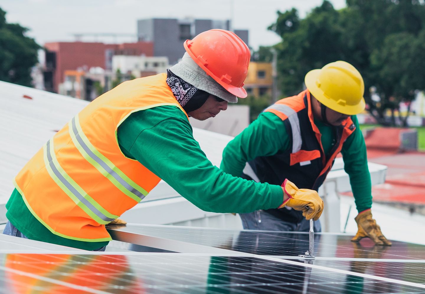 Two workers installing solar panels with buildings and a tree in the background.